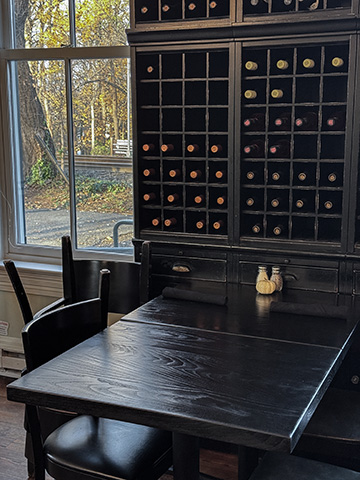 Blackened ash dining table in front of a tall built-in wine rack holding hundreds of bottles, with autumn light from a railside window
