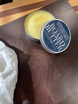 A tin of Kensington Woodworks Board Butter, open to show the beeswax inside, on a figured walnut surface